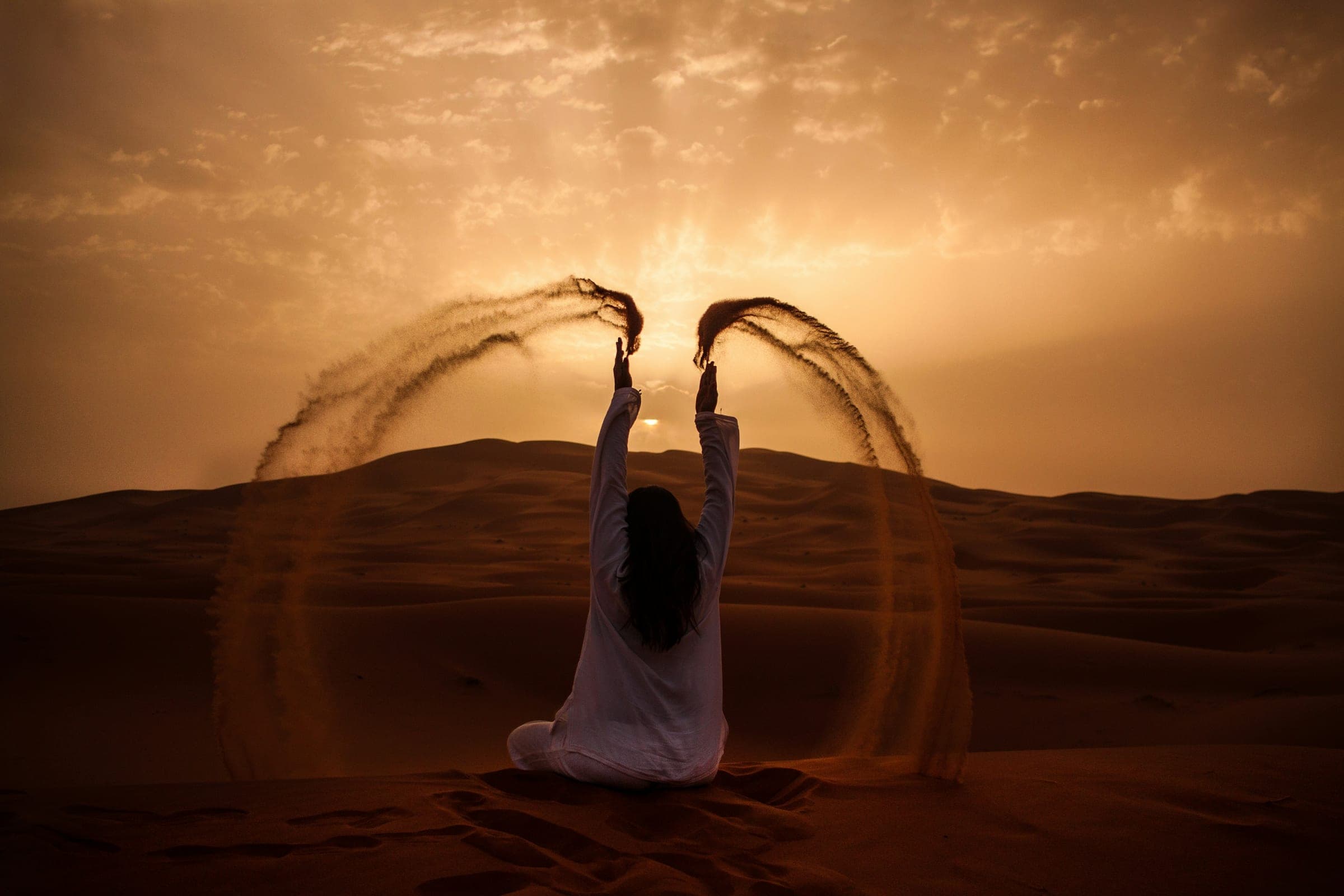 Een vrouw in het wit werpt bogen van warm Saharazand op tijdens zonsondergang